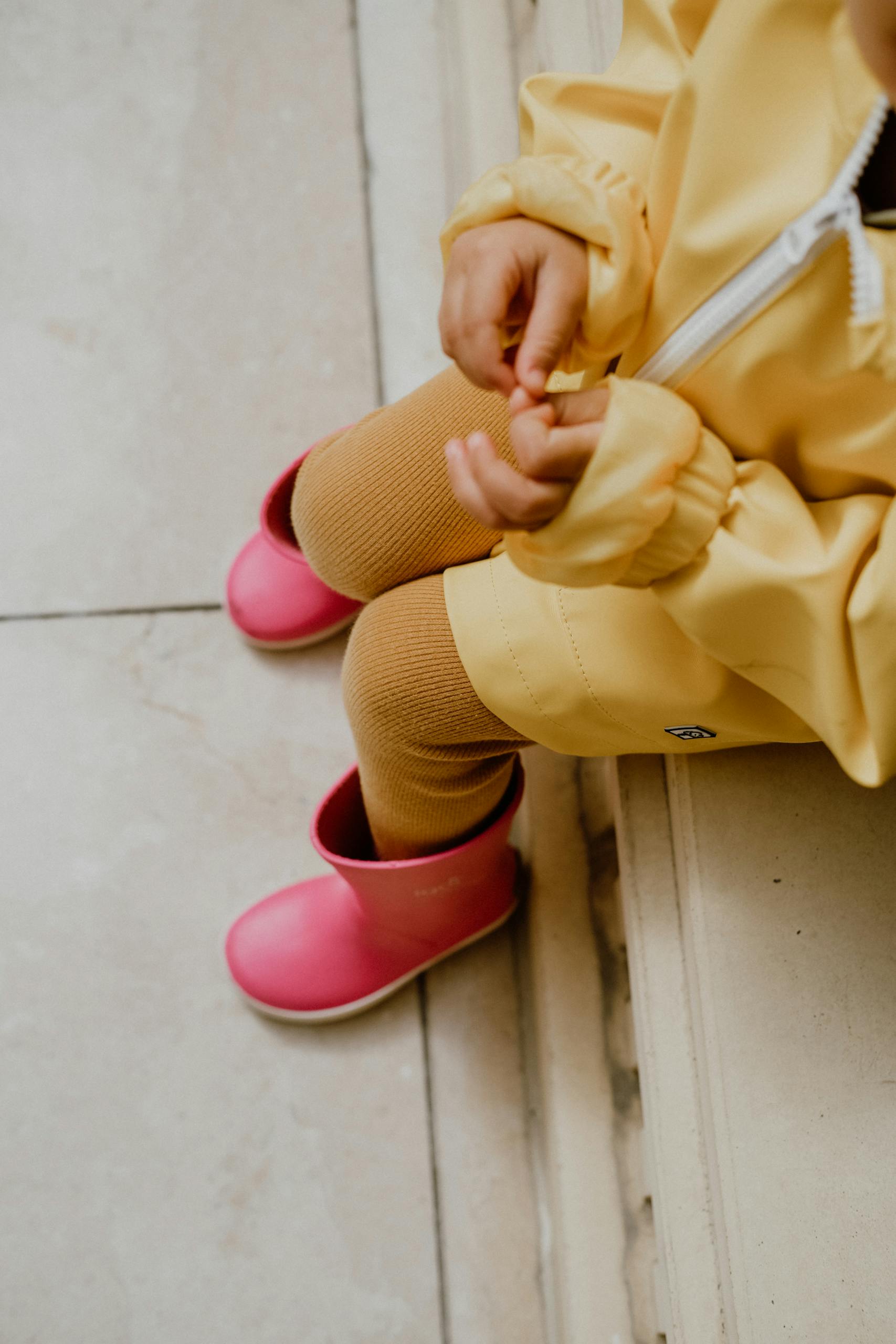 Top-down view of a child wearing a yellow raincoat and pink rain boots, sitting indoors.